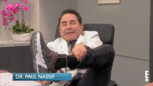 Dr paul Nassif taking of his shoe and revealing his socked foot on his desk